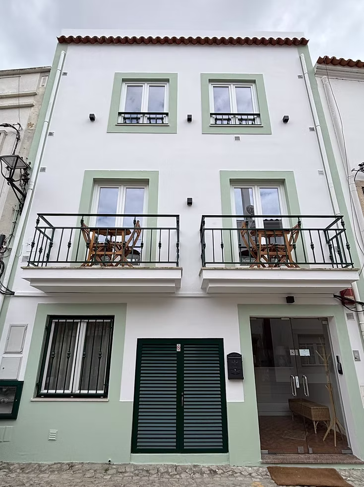 Prédio branco de três andares com detalhes em verde, duas varandas com cadeiras, janelas, portas duplas, caixas de correio e uma entrada com porta de vidro em uma rua de paralelepípedos.