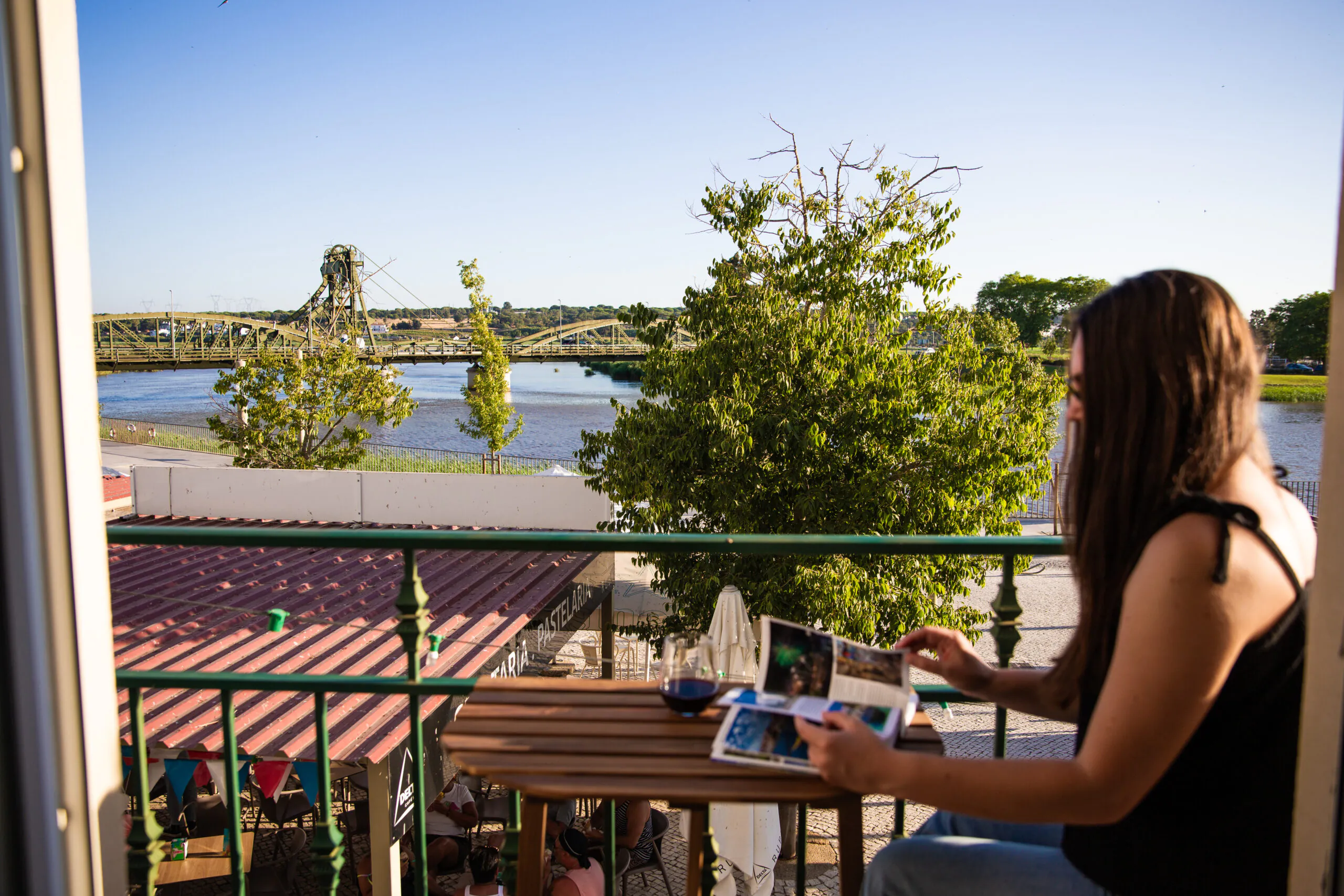 A person reads a magazine at a balcony table overlooking a river, bridge, and trees on a sunny day.