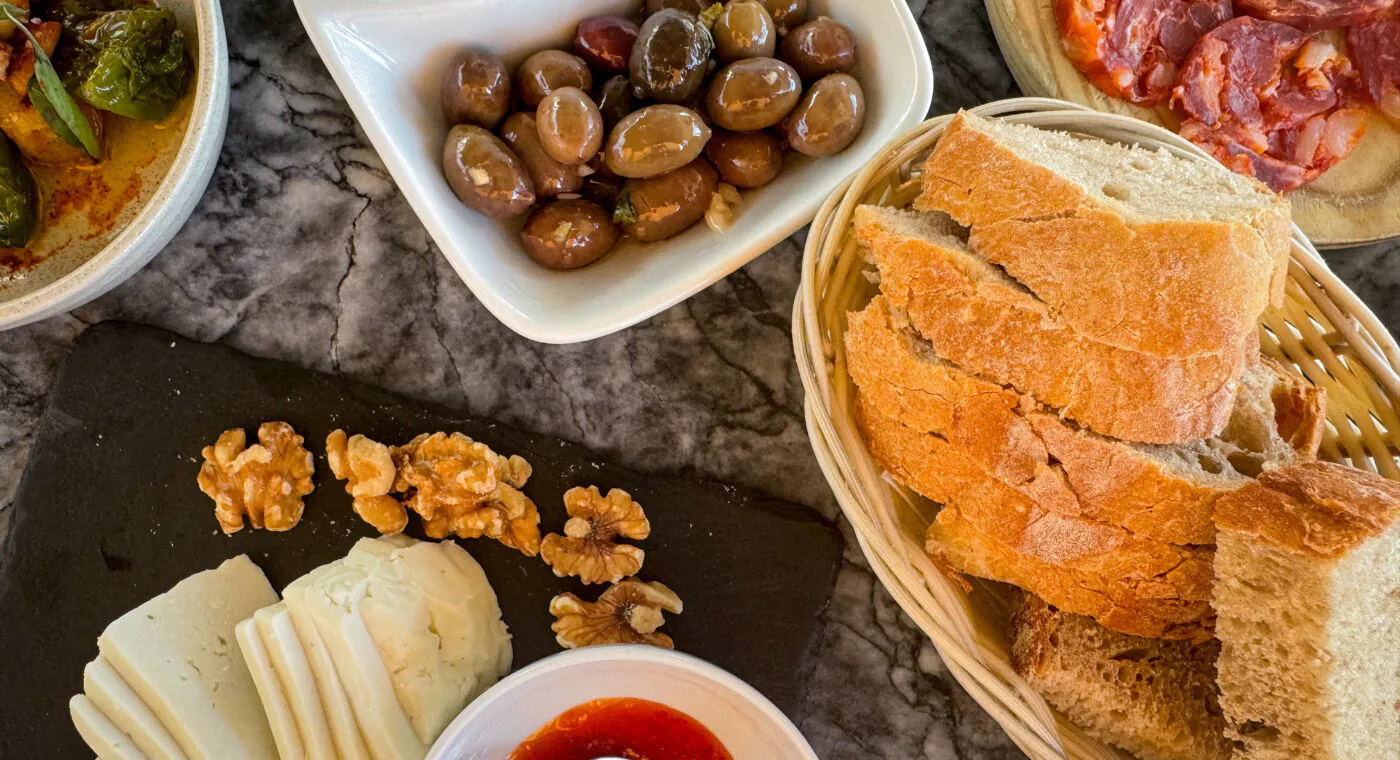Queijos variados, nozes, fatias de pão, azeitonas, chouriço e molho de tomate dispostos sobre uma mesa de mármore, vista de cima.