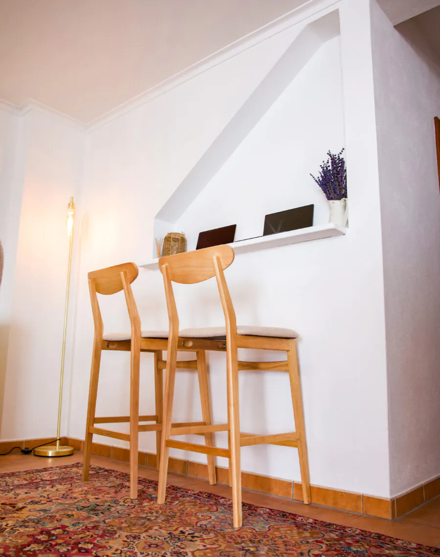 Two wooden bar stools at a white counter with a vase of lavender and small items, on a patterned rug in a cozy, well-lit room.