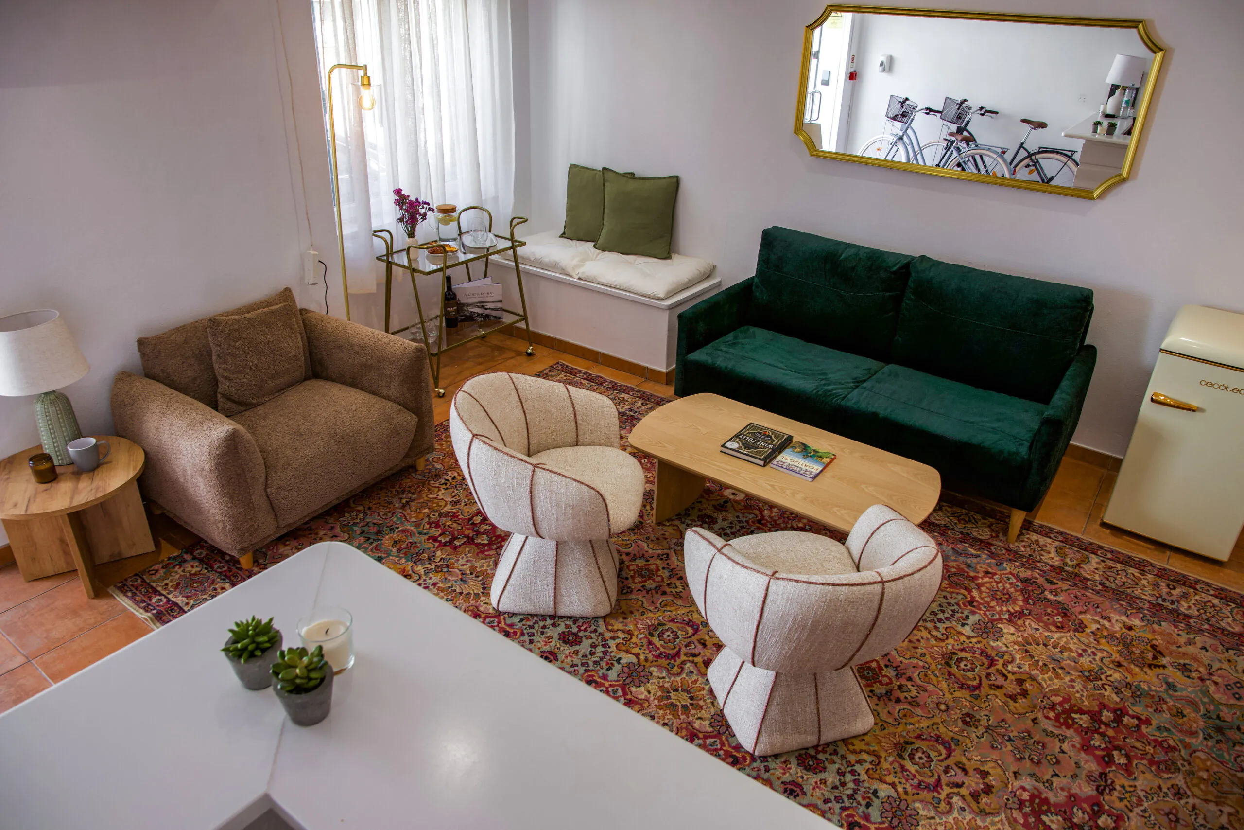 Cozy living room with green sofa, two round chairs, armchair, coffee table, vintage rug, gold mirror, mini fridge, and a bicycle seen in the mirror.