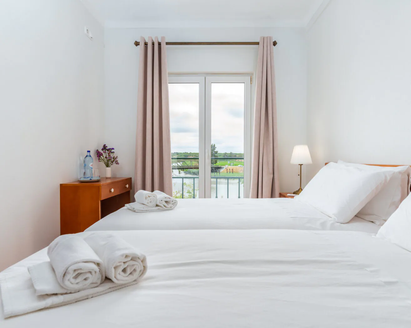 A bright, tidy bedroom with two twin beds, white linens, a window with beige curtains, and a view of greenery outside.