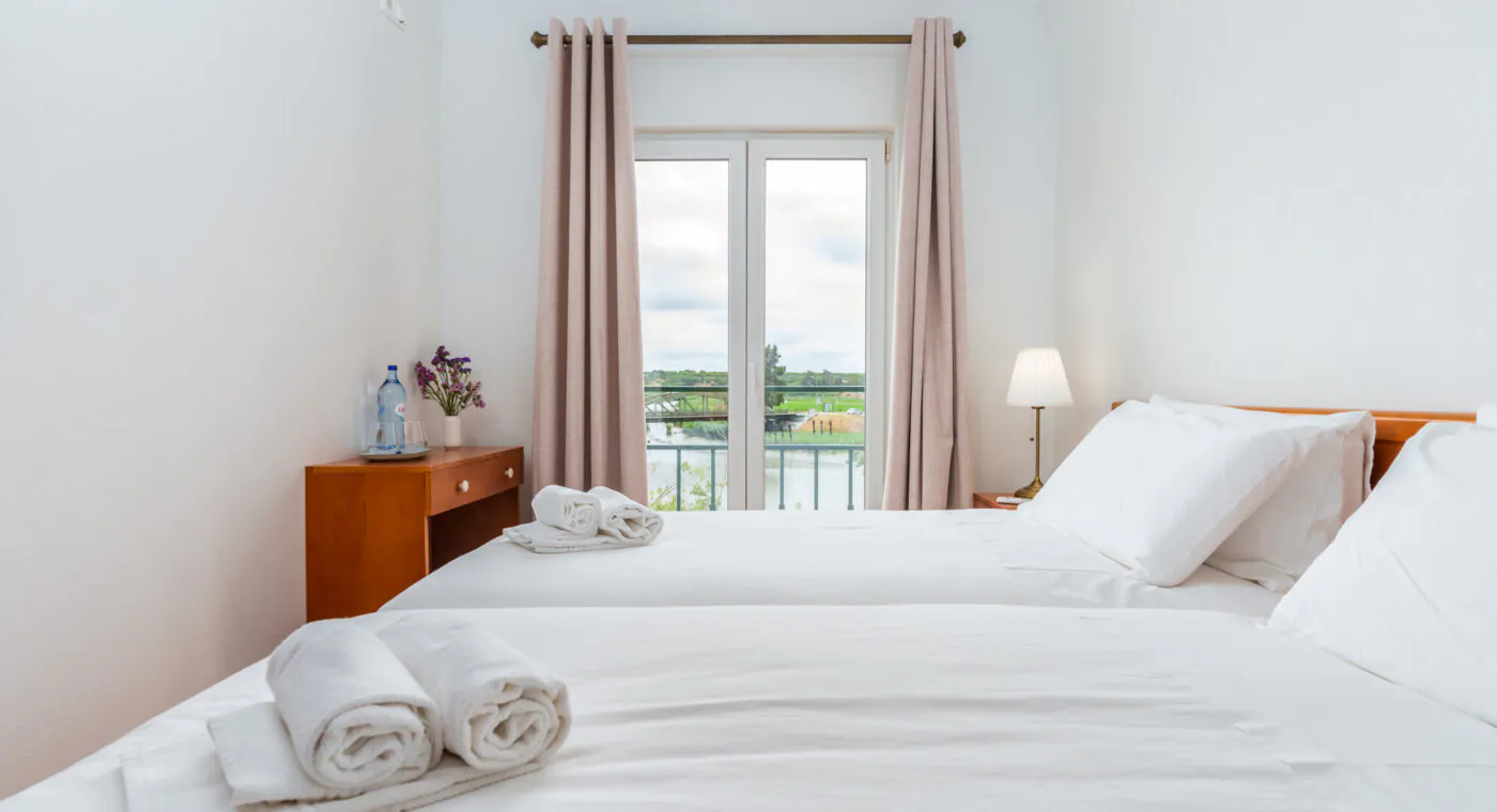 A bright, tidy bedroom with two twin beds, white linens, a window with beige curtains, and a view of greenery outside.