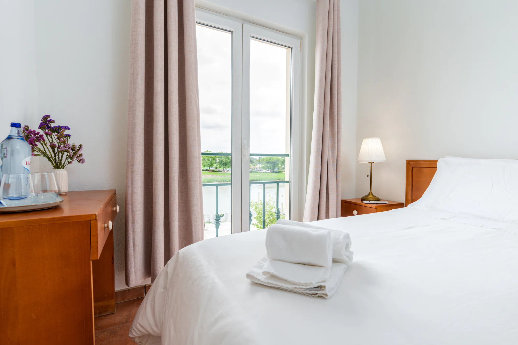 Bright bedroom with a neatly made bed, folded towels, wooden furniture, lamp, flowers, and a window overlooking greenery and water.