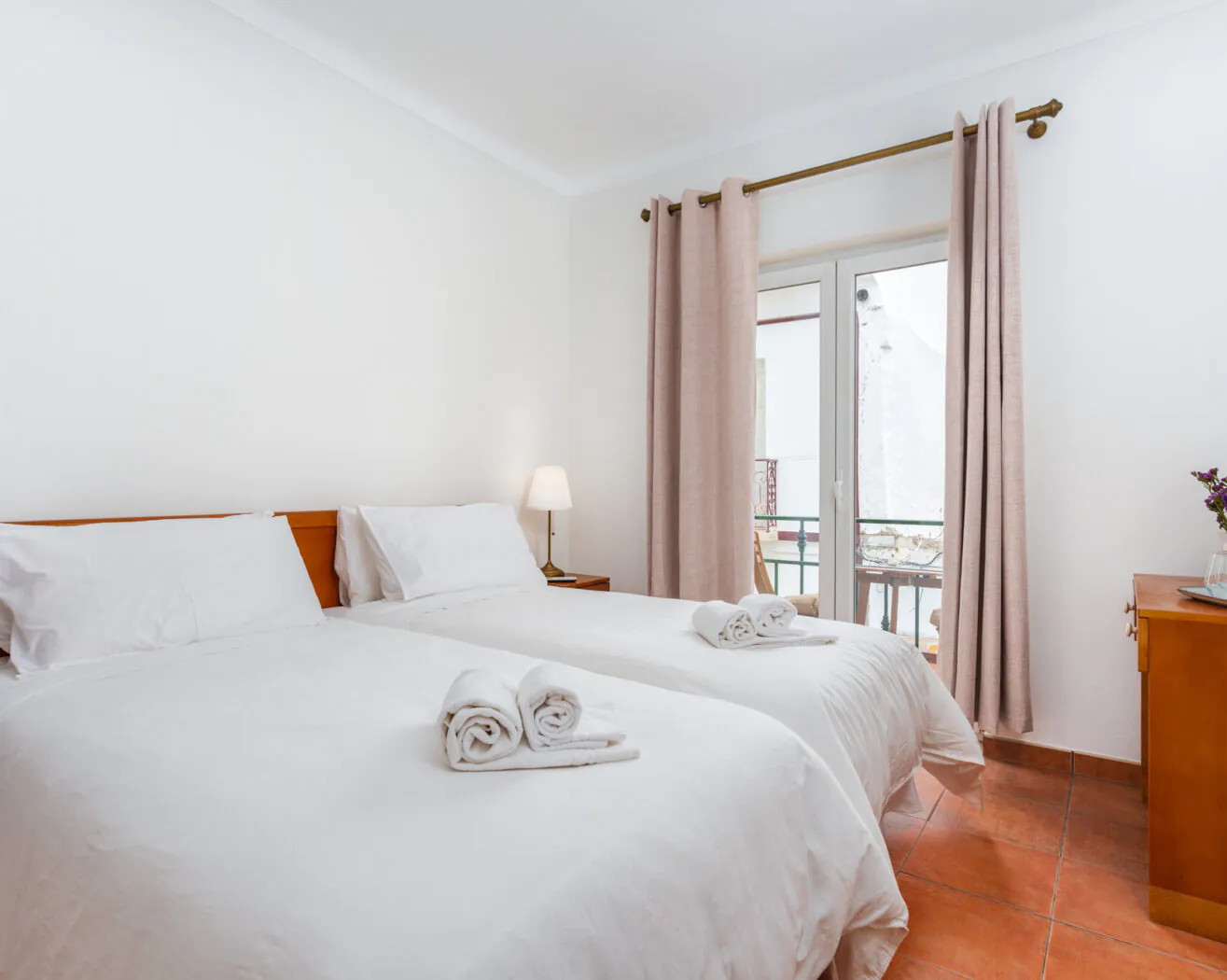 Bright bedroom with two single beds, white linens, a lamp, window with beige curtains, and a small dresser with flowers and water bottle.