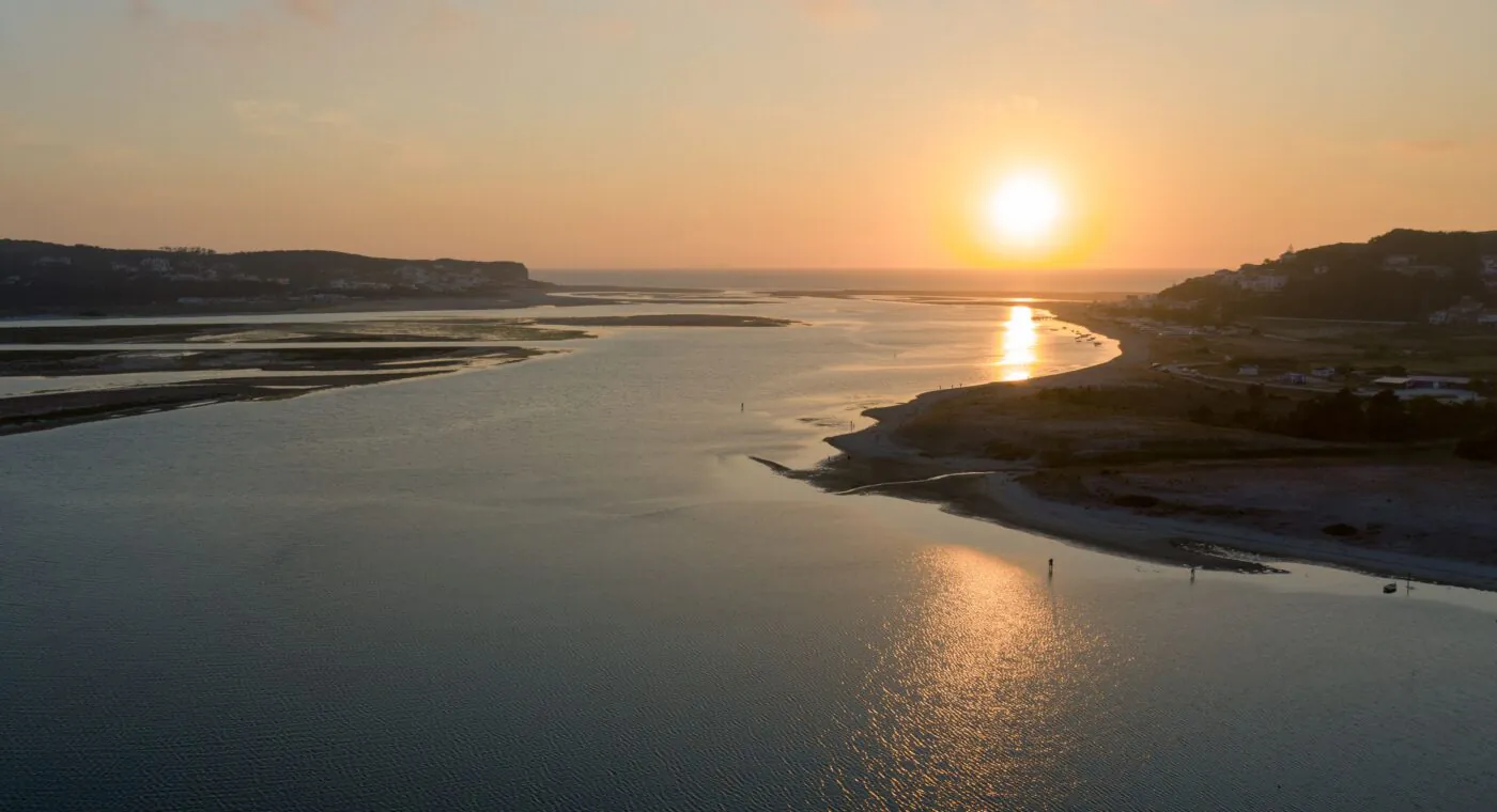 Um sereno estuário de rio ao pôr do sol, com ondas suaves e o sol projetando um reflexo dourado na água.