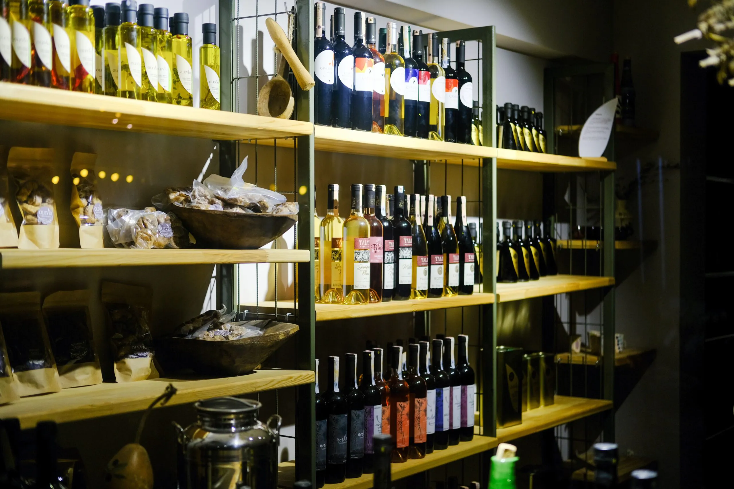 Garrafas de vinho e azeite estão dispostas em prateleiras de madeira ao lado de potes, sacos e tigelas com alimentos em uma loja com iluminação aconchegante.