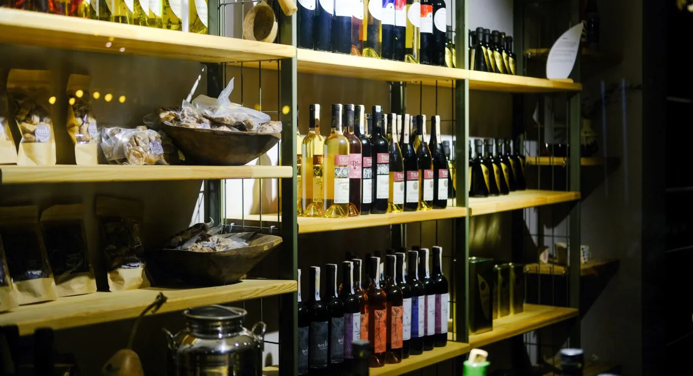 Bottles of wine and olive oil are displayed on wooden shelves, with packages of snacks and jars beside them in a softly lit store.