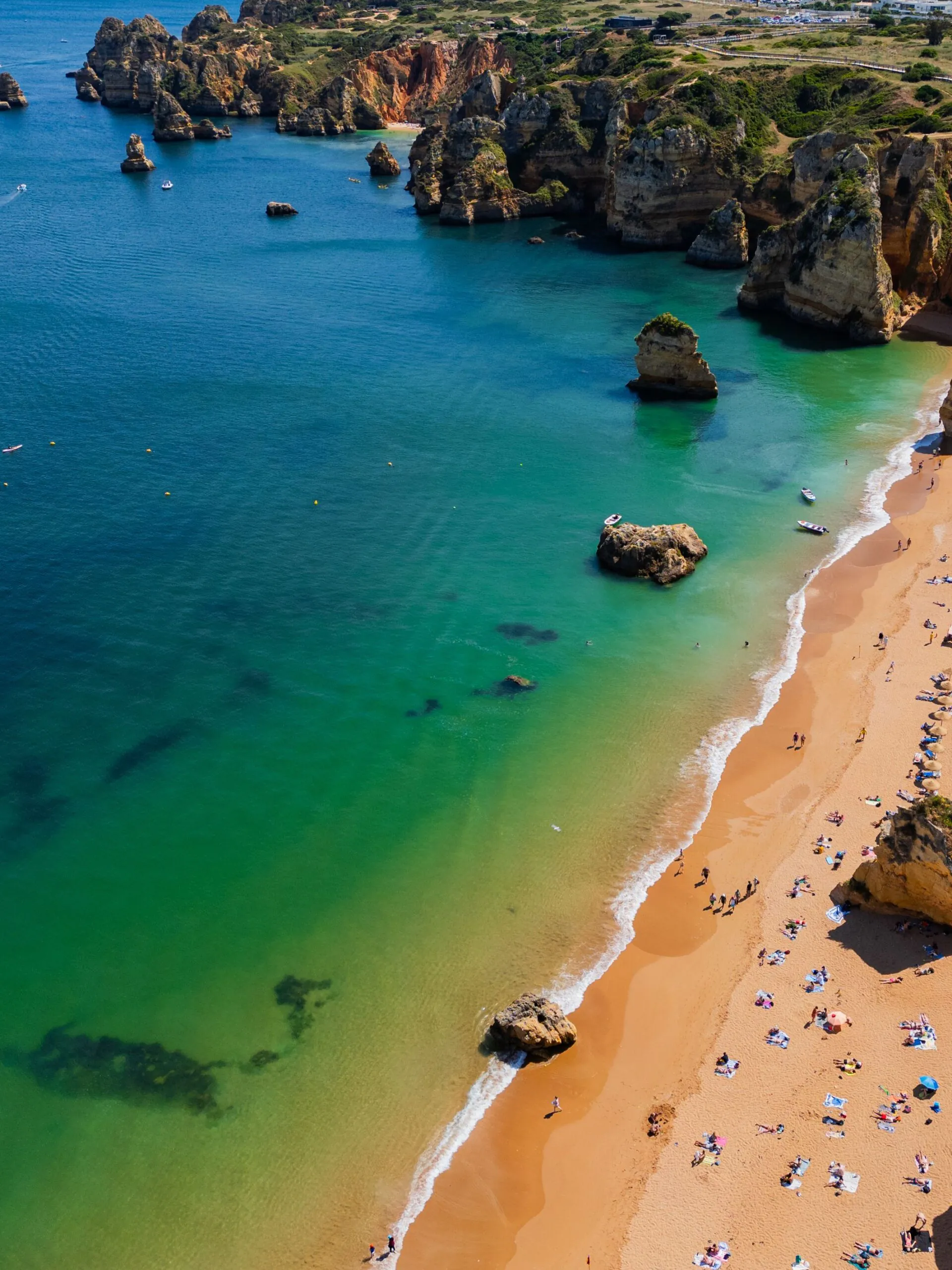 Aerial view of a sandy beach with sunbathers, turquoise water, and rocky cliffs along the coastline.