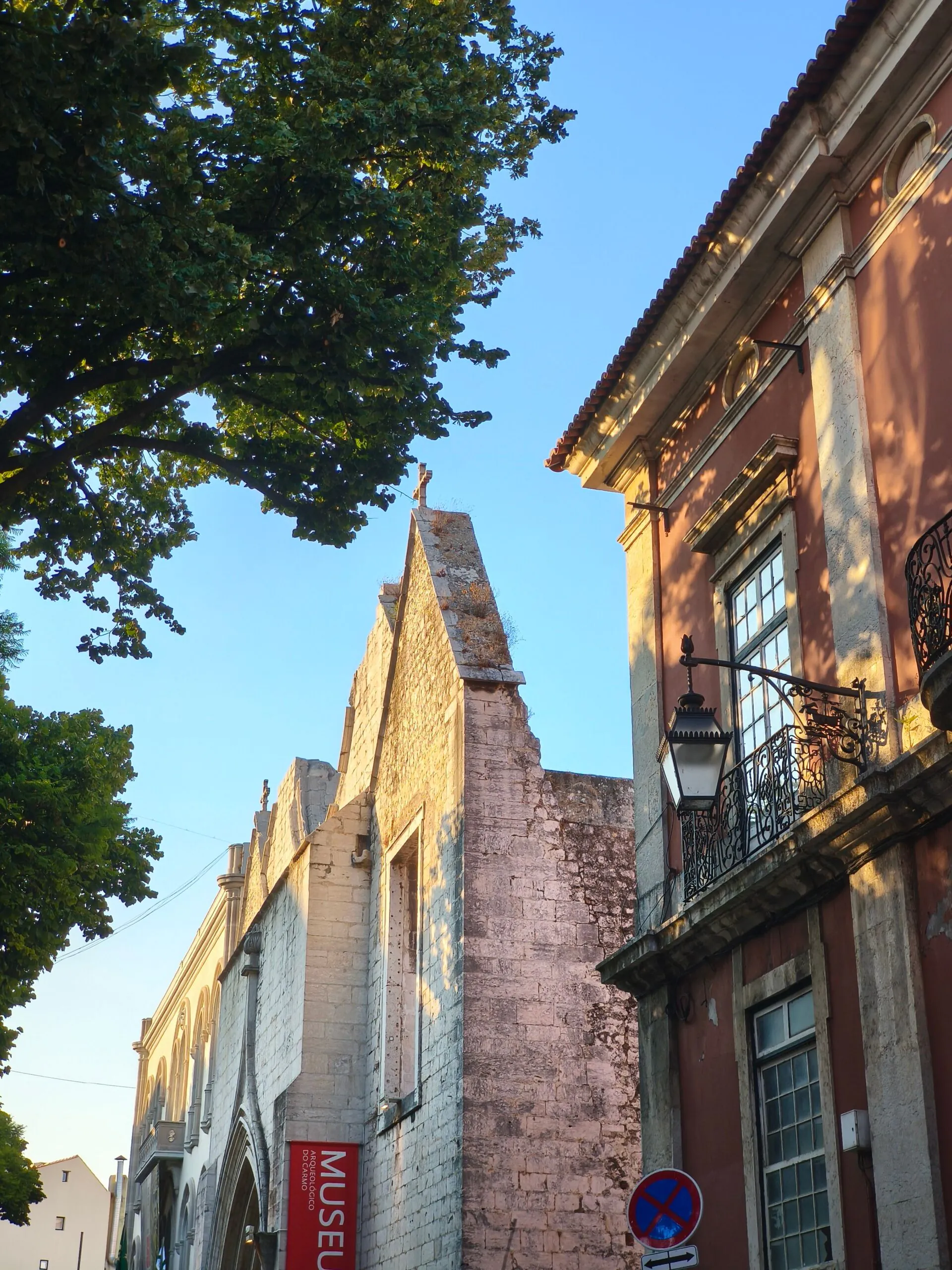 Sunlit historic stone and brick buildings with balconies and a museum sign, tree branches overhead, and a no parking sign in the foreground.
