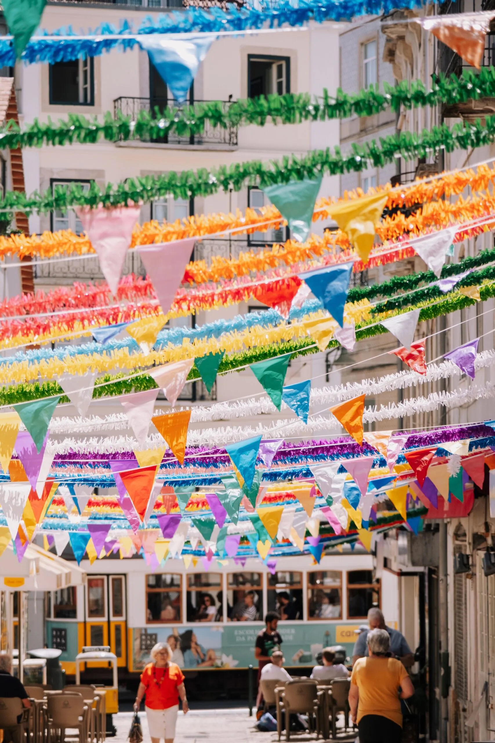 Colorful bunting and streamers hang above a bustling street with people walking and a tram passing by in a European city.