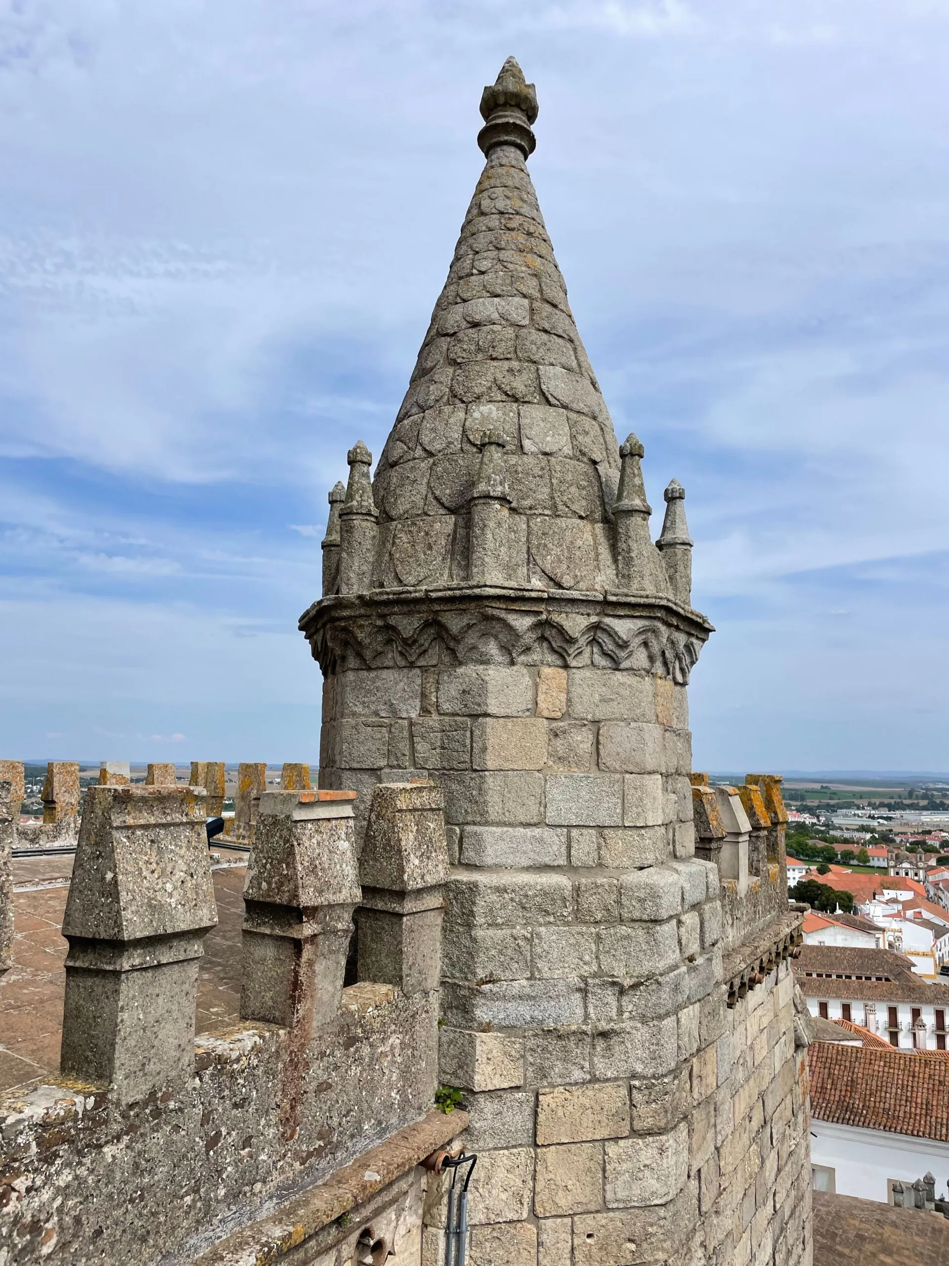Stone turret with a conical roof and decorative battlements overlooks a town with white buildings under a partly cloudy sky.