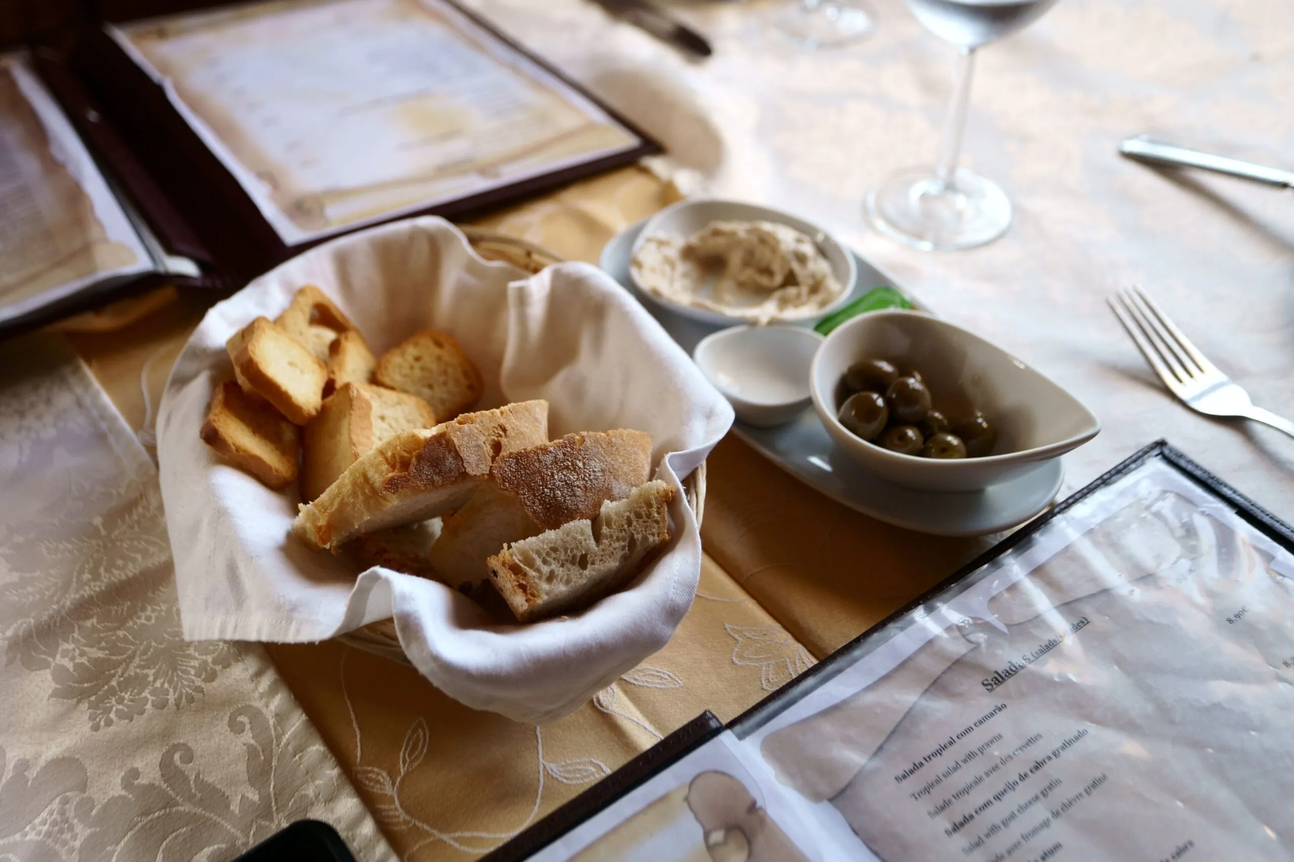 Uma cesta de pão fatiado com um prato de azeitonas verdes e molho sobre uma mesa posta com cardápios, talheres e uma taça de vinho.