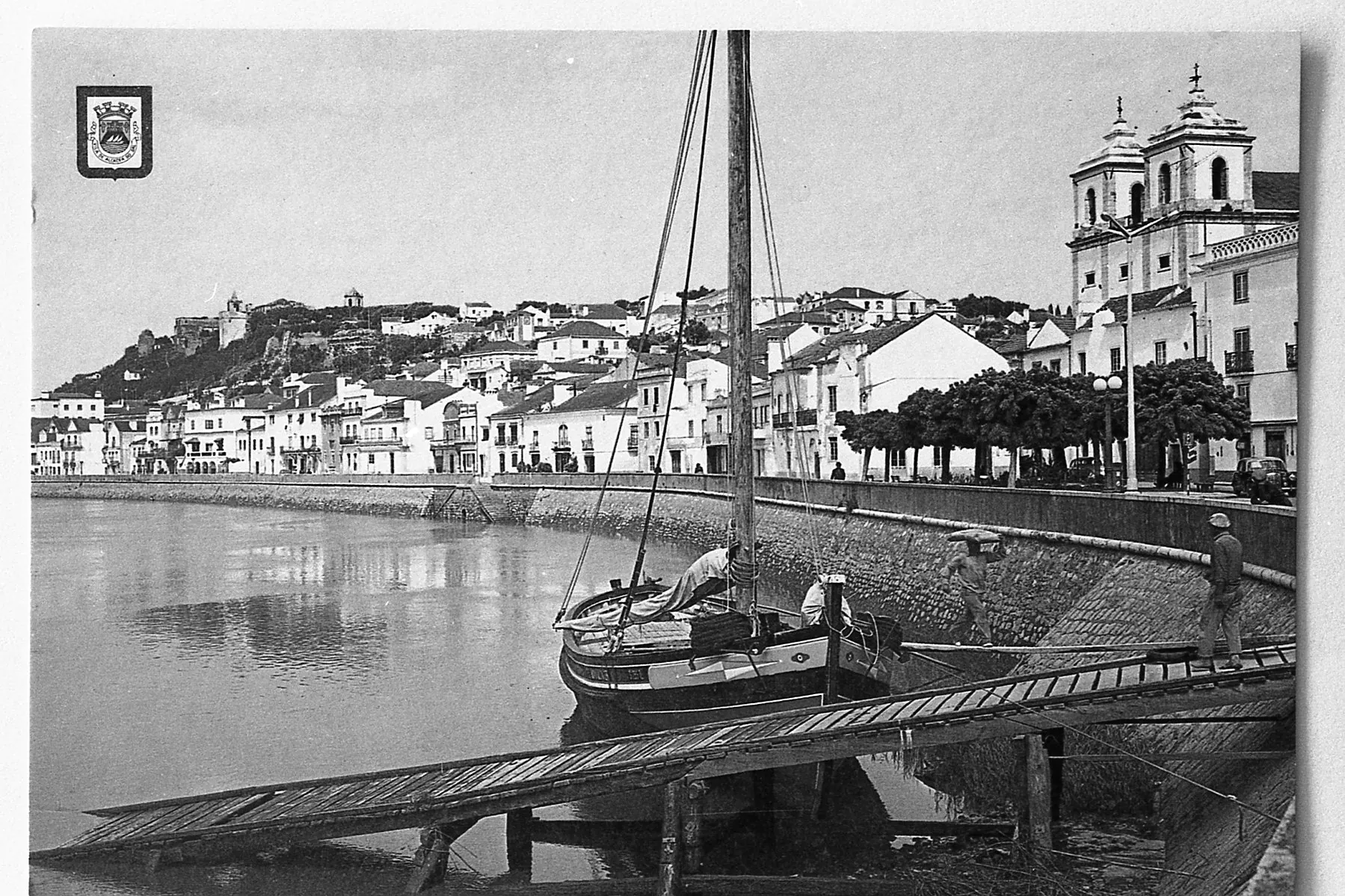 Um pequeno veleiro atracado em uma cidade ribeirinha com prédios brancos, uma igreja e um castelo no alto de uma colina ao longe. Duas pessoas podem ser vistas perto do barco.
