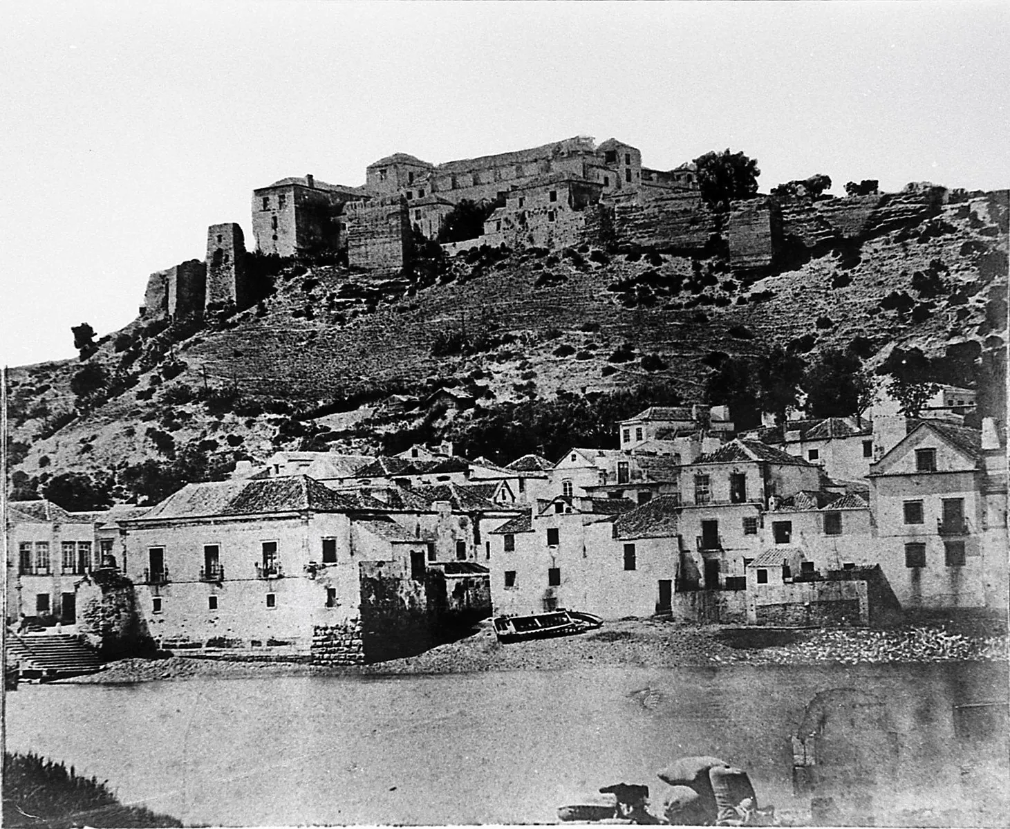 Foto em preto e branco de casas no sopé de uma colina, no topo da qual se ergue uma grande fortaleza ou castelo antigo, com árvores espalhadas e terreno acidentado.