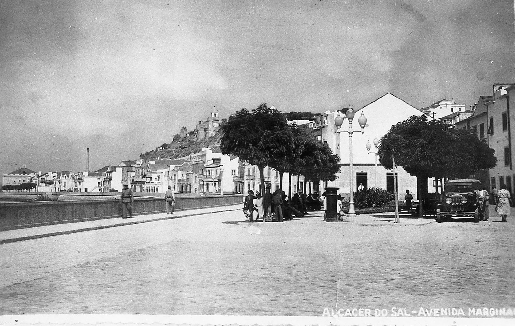 Black and white photo of Alcácer do Sal’s riverside avenue, with people, vintage cars, trees, and historic buildings along the waterfront.